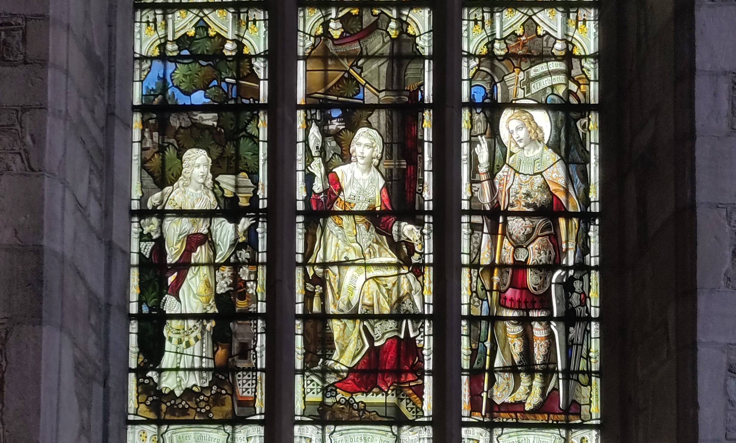 A photograph of stained glass depicting three figures, one in each light. The leftmost figure is a young girl, the middle figure a woman spinning thread and the rightmost figure is a depcition of St Michael in armour carrying a shield and standard. He has wings which are made of peacock feathers.